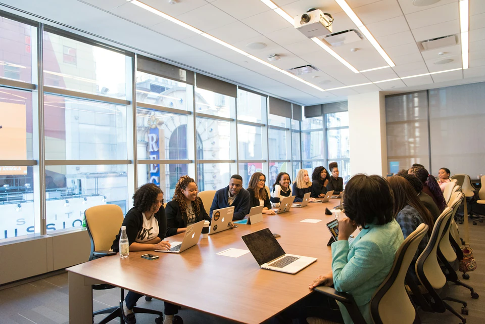 Group of people sitting beside rectangular wooden table with laptops.